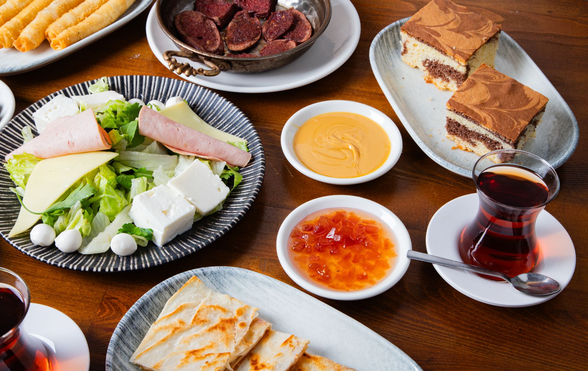 Ein reich gedeckter Tisch mit verschiedenen süßen Speisen, Kuchen und traditionellem türkischen Tee in Gläsern auf dunklem Holz.