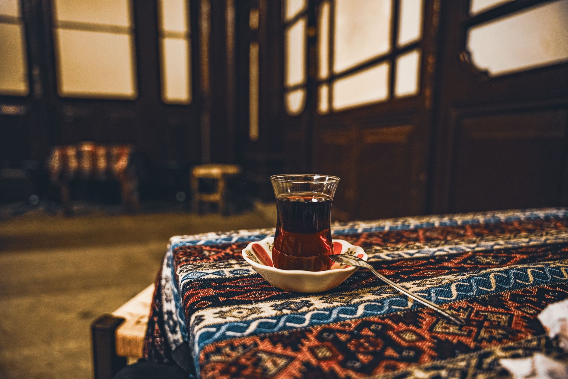 Ein traditionelles türkisches Teeglas mit rötlich schimmerndem Tee auf einer gemusterten Tischdecke in einem gemütlich beleuchteten Innenraum.